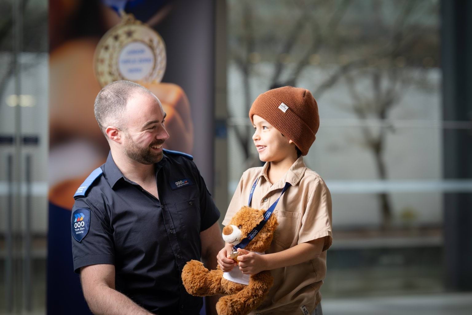 A male Triple Zero Call-Taker in uniform kneeling next to his young male hero he nominated who is holding Benny the Bear