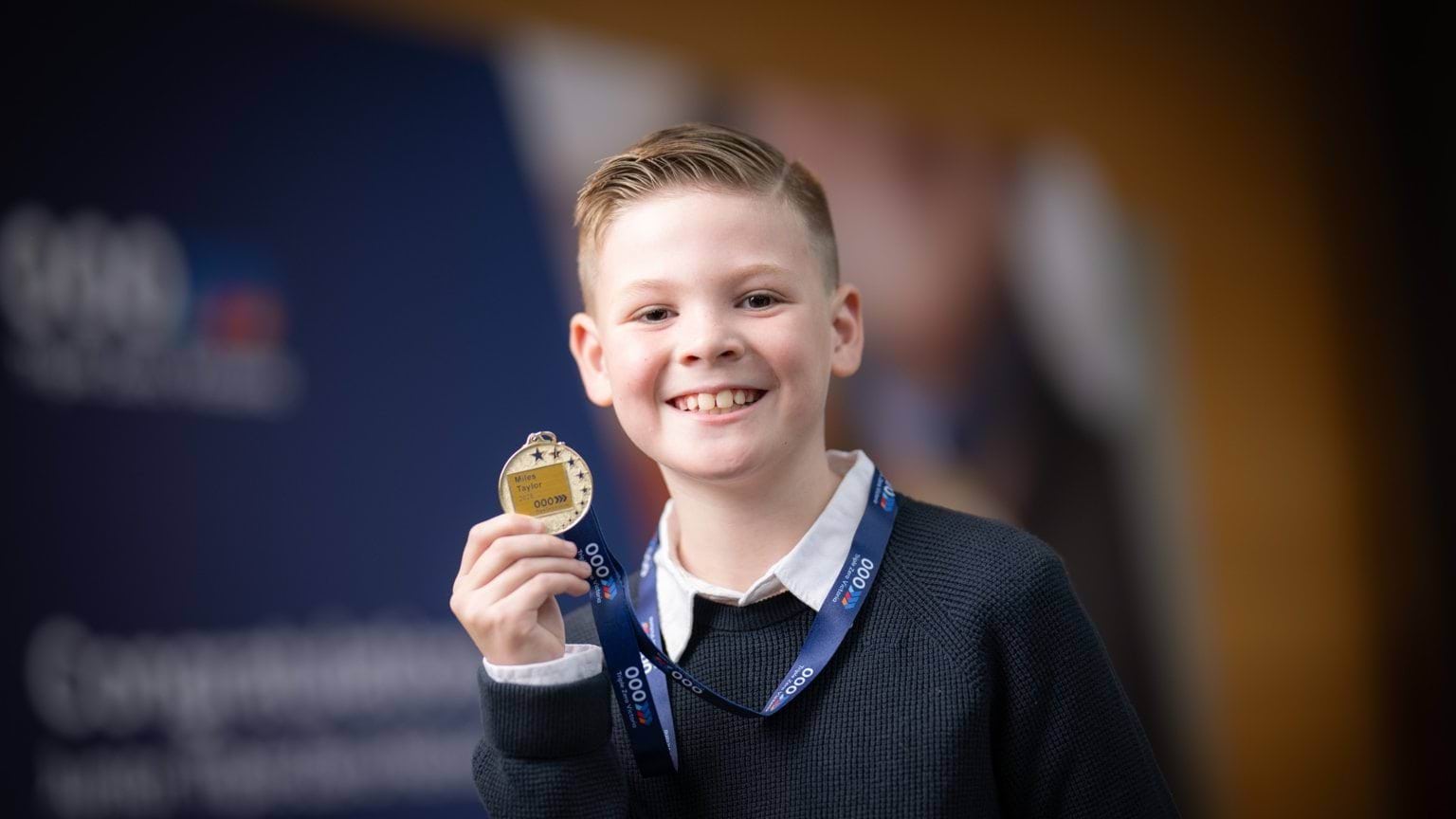 A young boy smiling holding up his medal at the camera