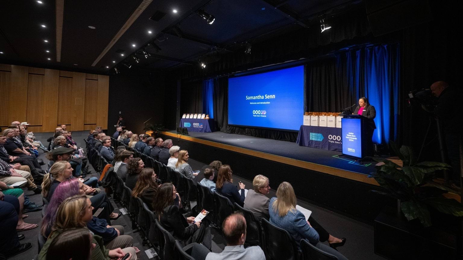 A theatre style row crowd sits in front of a stage with a presenter and large screen as part of the official presentation.